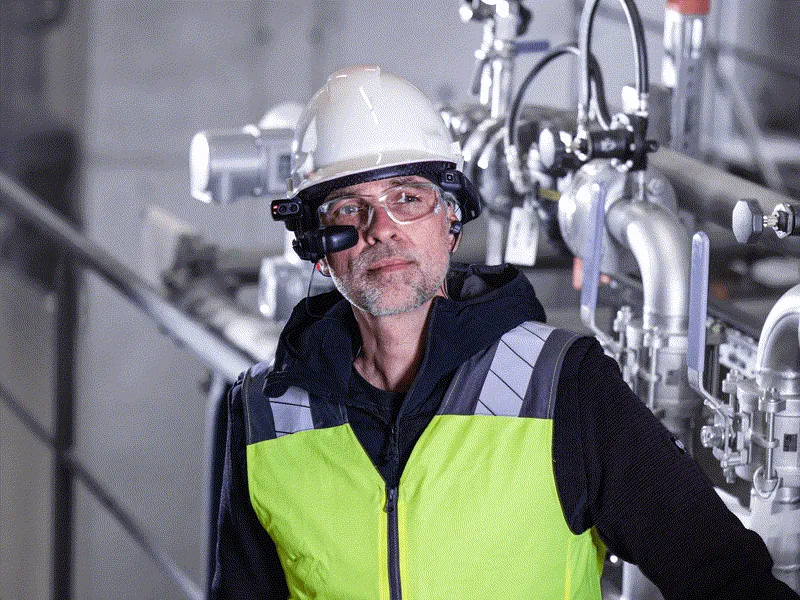 A technician in a white hard hat wearing clear safety glasses with a helmet-mounted heads-up display in front of them, in a plant room